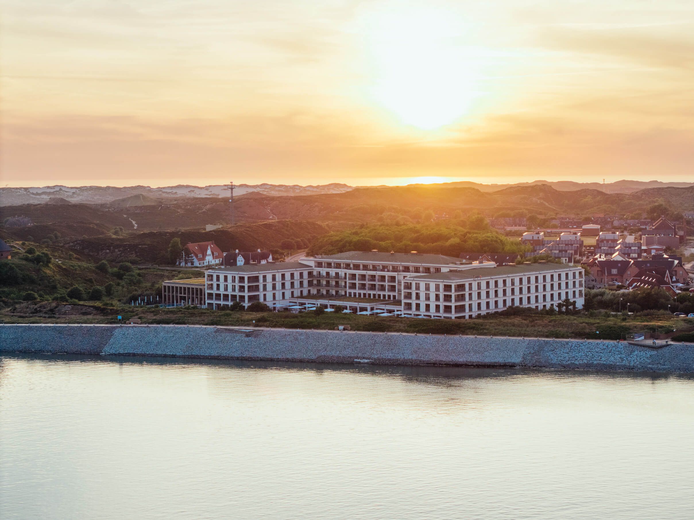 Ein Sonnenuntergang über einem modernen Hotel am Wasser, umgeben von sanften Hügeln und ruhigen Häusern.