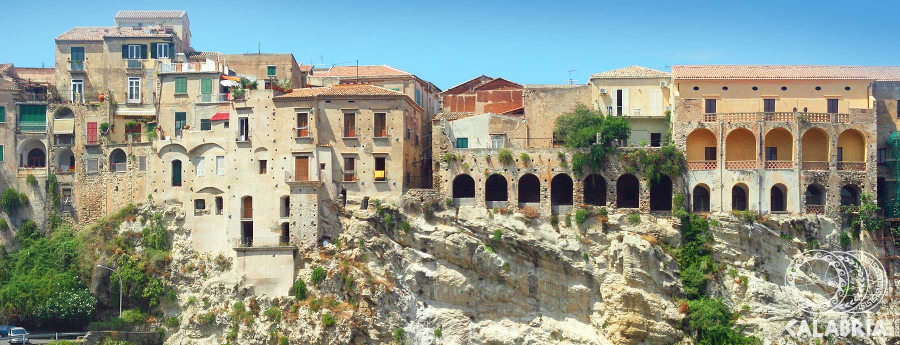 Eine alte Stadt mit historischen Gebäuden auf einem Felsen. Üppige Vegetation und blauer Himmel prägen die Szenerie.