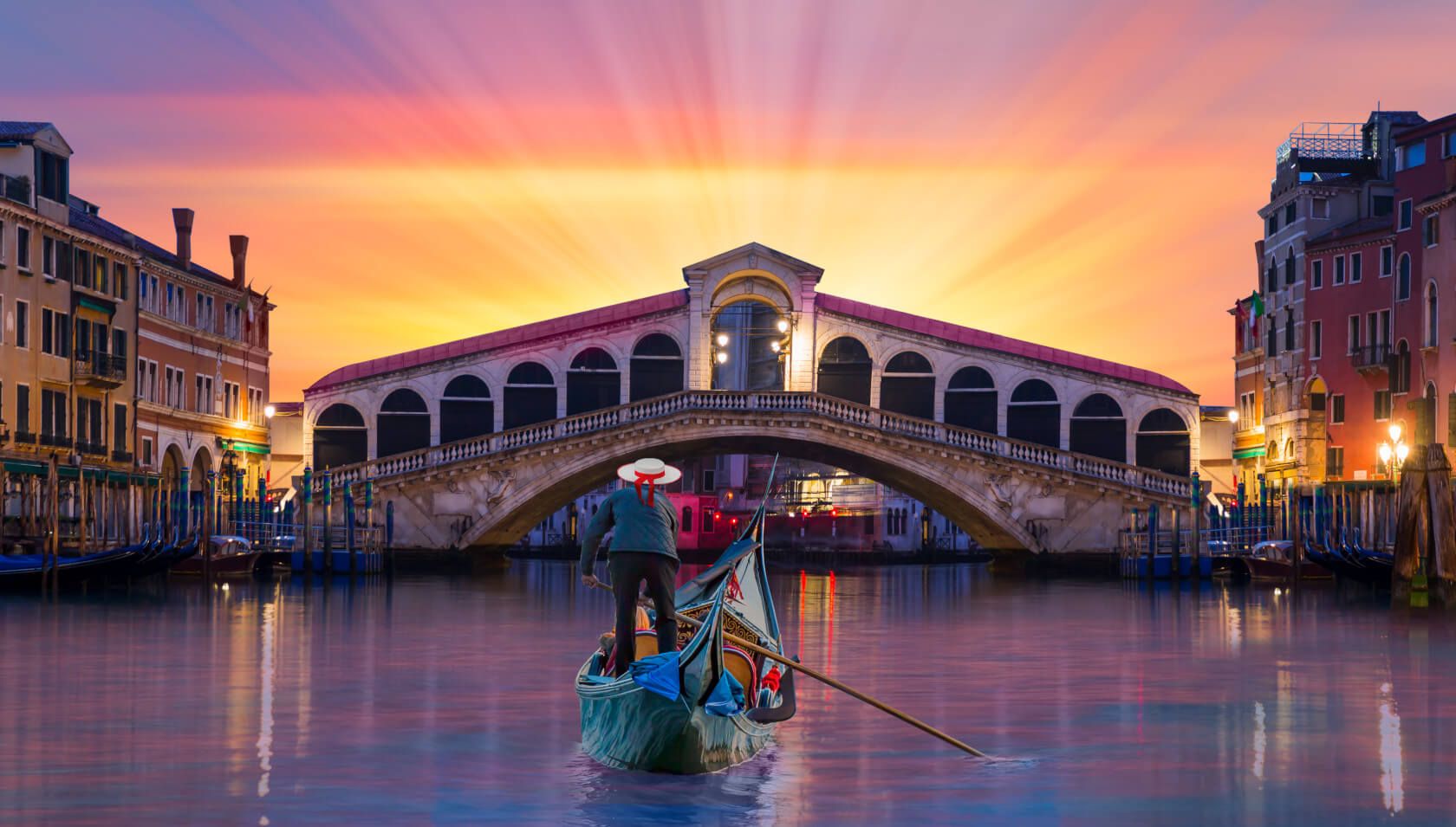 Ein Gondoliere fährt bei Sonnenuntergang unter der Rialtobrücke in Venedig. Die Farben sind lebhaft und malerisch.