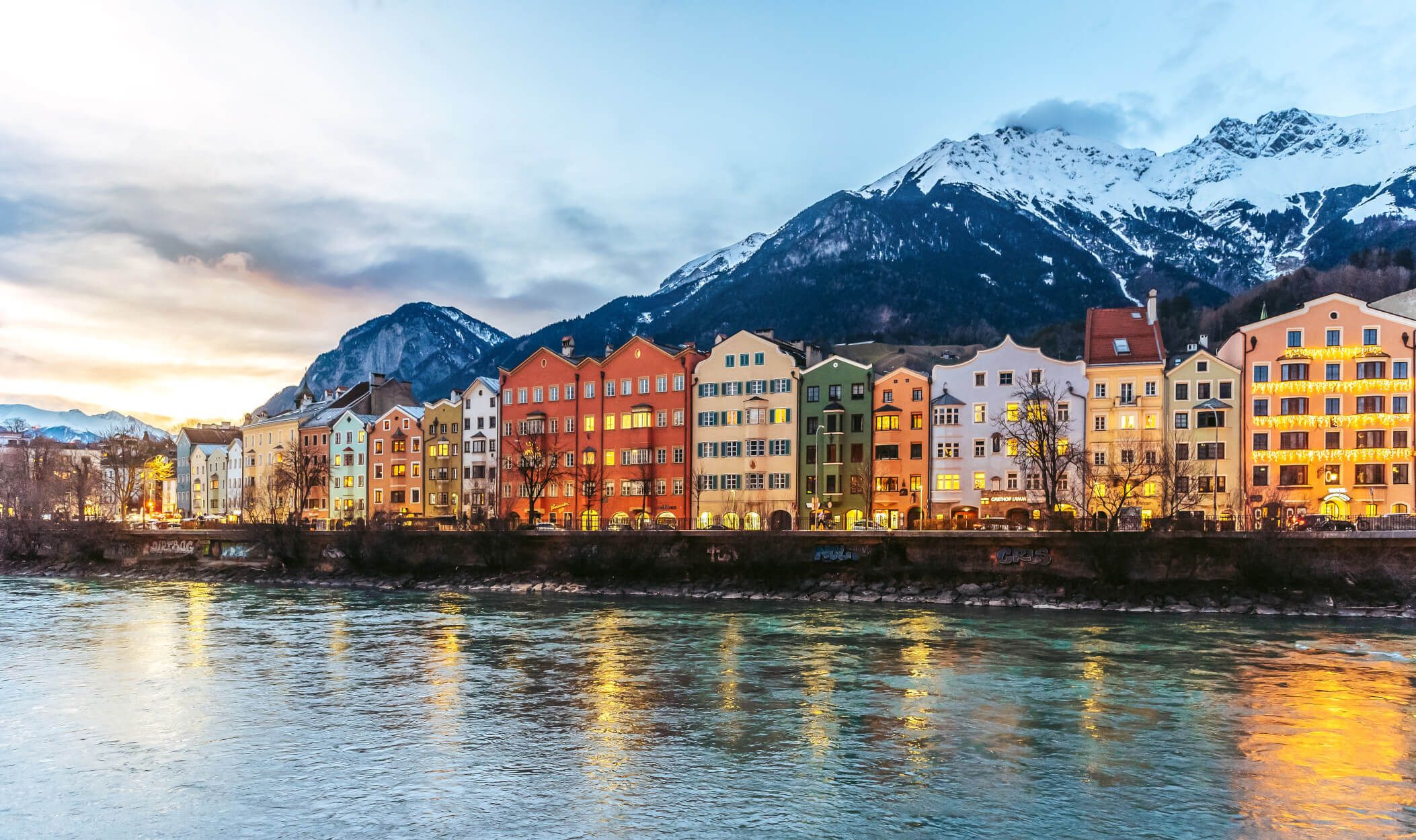 Bunte Häuser am Ufer eines Flusses, umgeben von schneebedeckten Bergen im Abendlicht. Schöne alpine Landschaft.