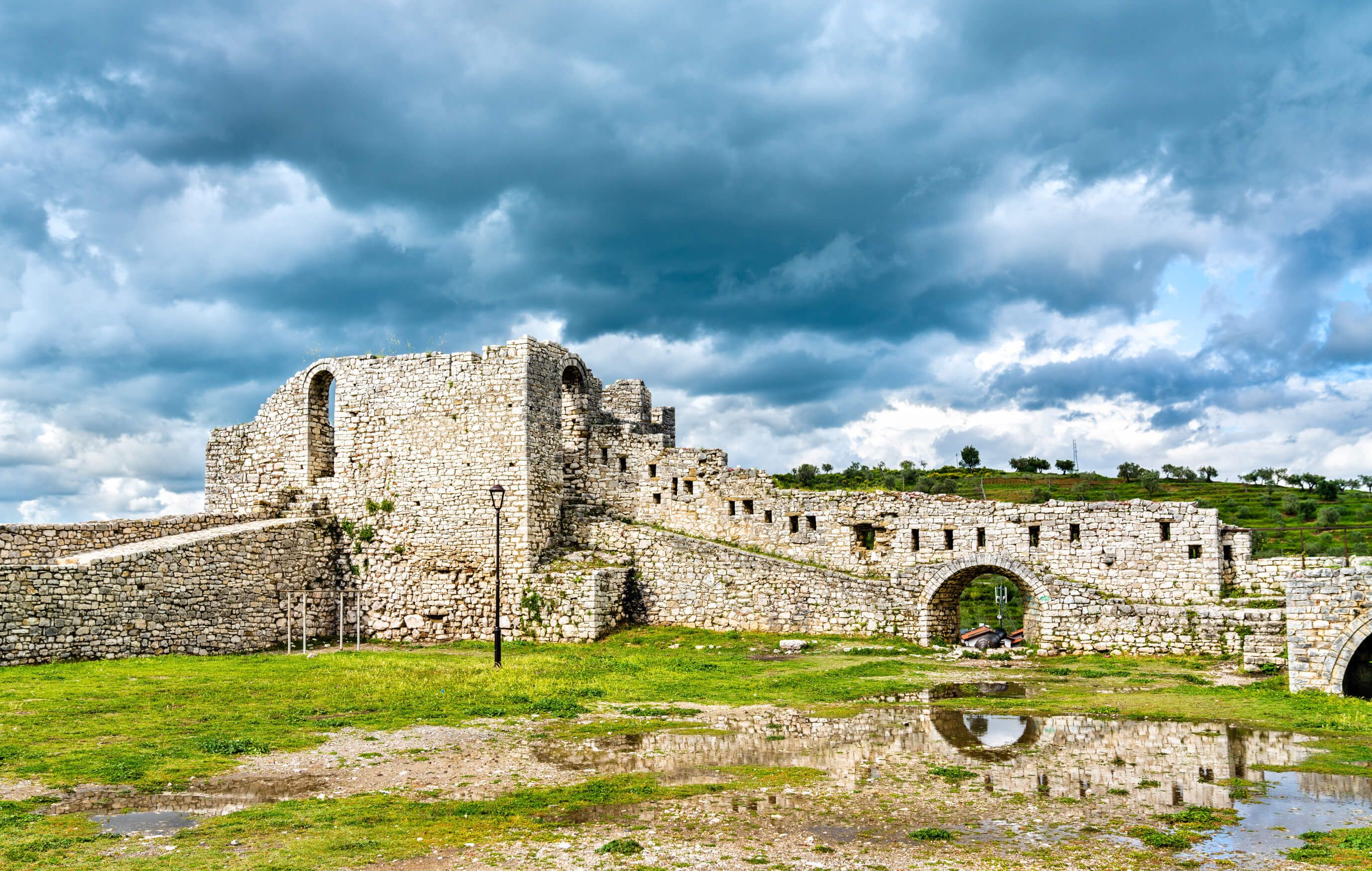 Ruine eines alten steinernen Bauwerks, umgeben von Wiese und Schatten durch dunkle Wolken im Hintergrund.