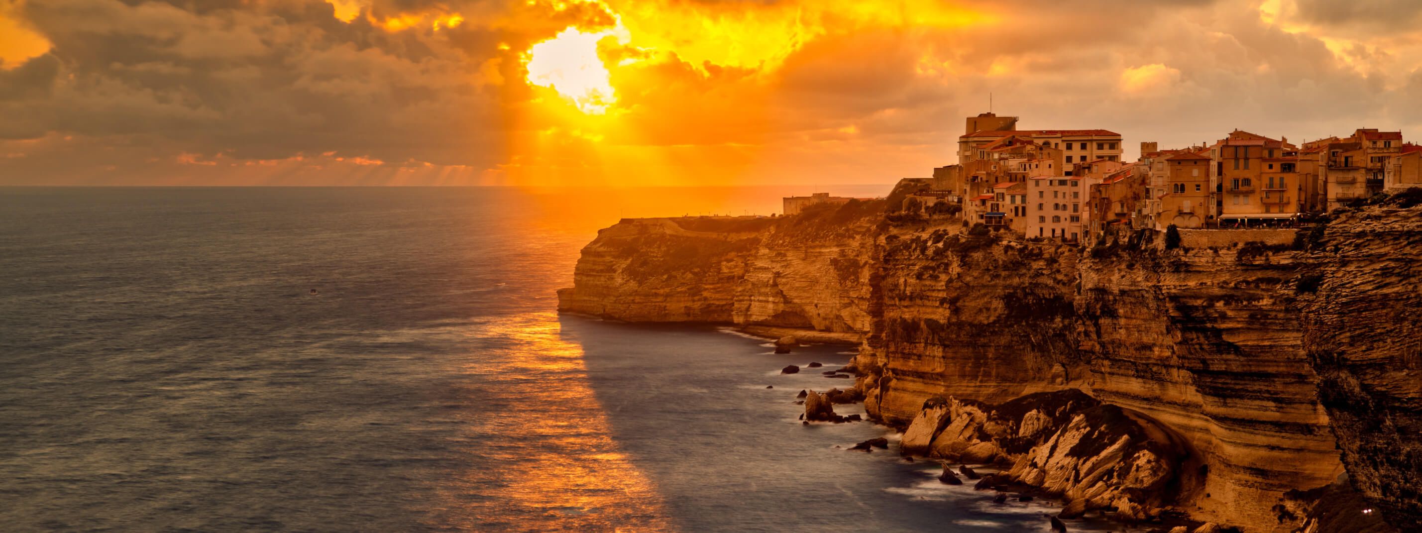 Ein Sonnenuntergang über einem Küstenort, umgeben von steilen Klippen und glitzerndem Wasser.