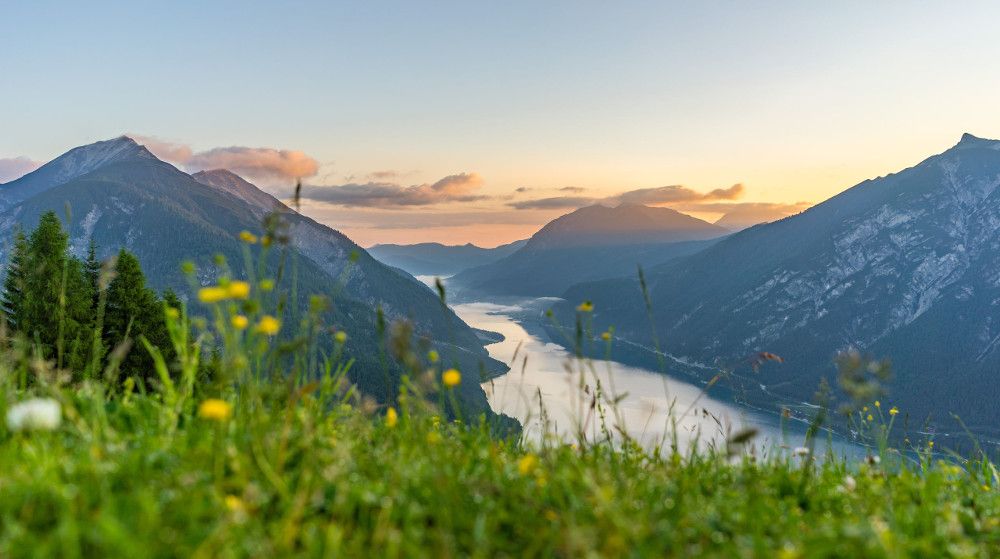 Malerische Berglandschaft bei Sonnenuntergang, mit einem ruhigen Fluss und blühenden Wiesen im Vordergrund.