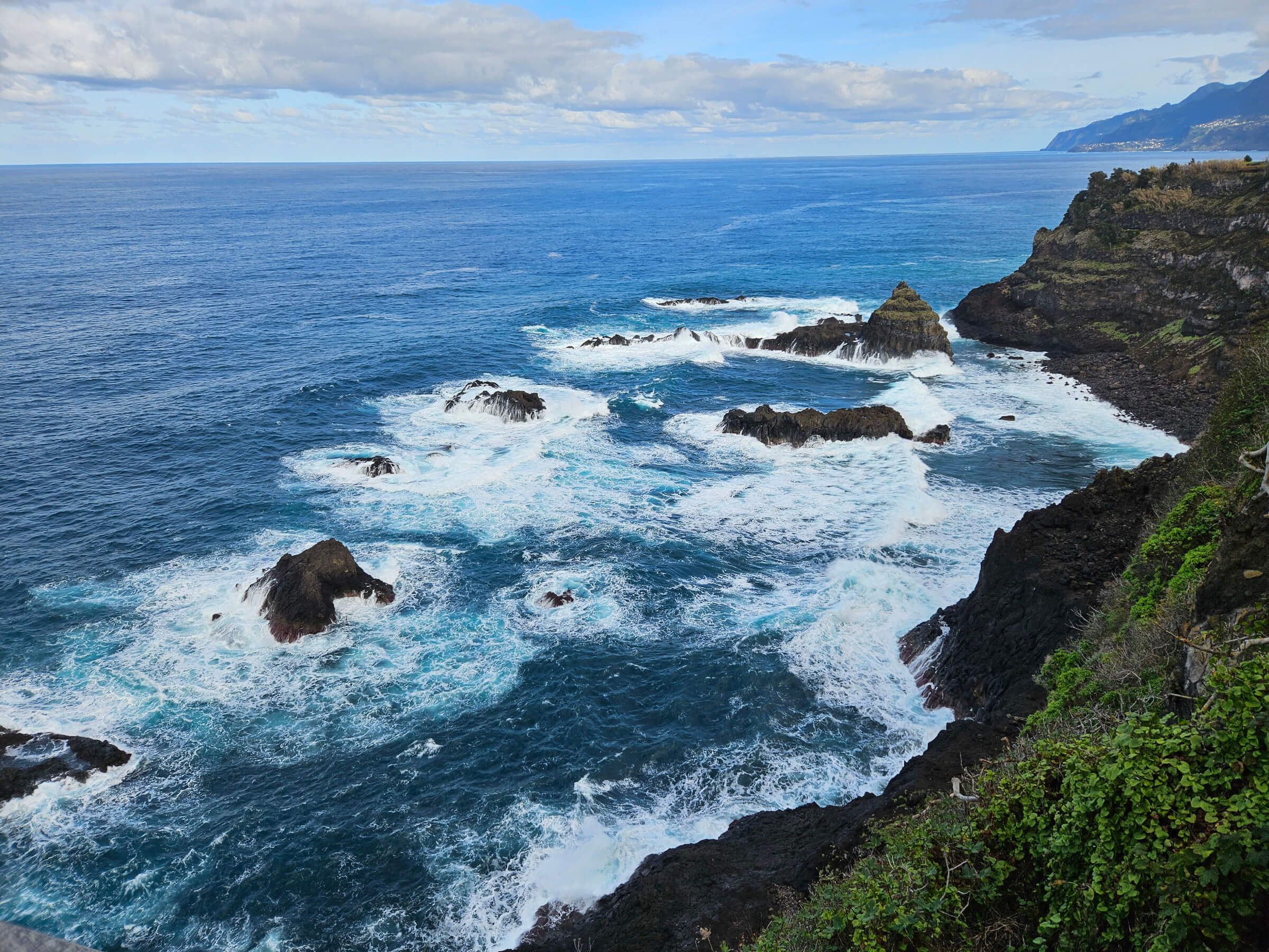 Die Küste zeigt felsige Klippen, sanfte Wellen und einen weiten blauen Ozean unter einem bewölkten Himmel.